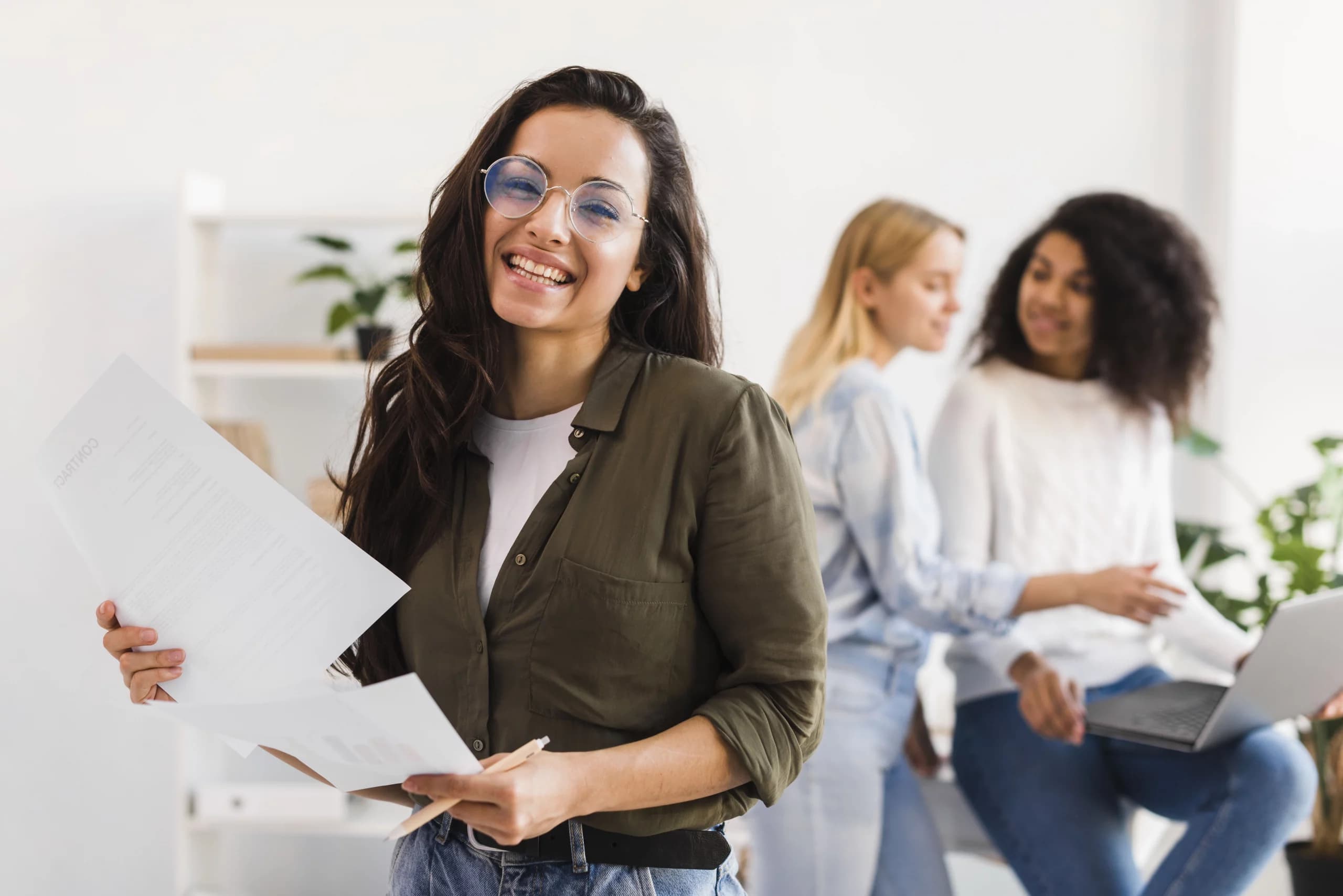 Jeune femme souriante tenant des documents, avec deux collègues travaillant en arrière-plan.