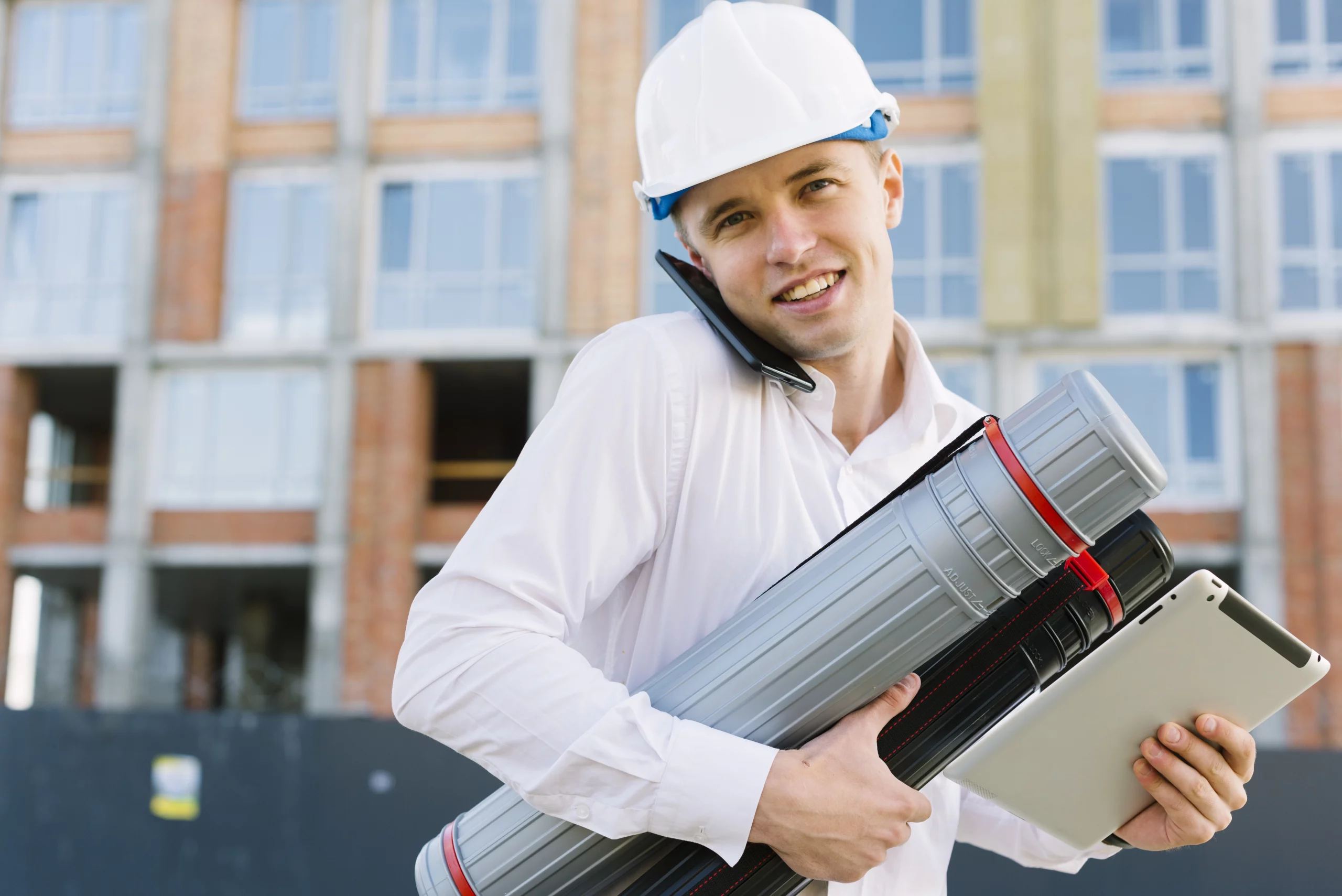 Conducteur de travaux souriant au téléphone sur un chantier, portant des plans et une tablette.